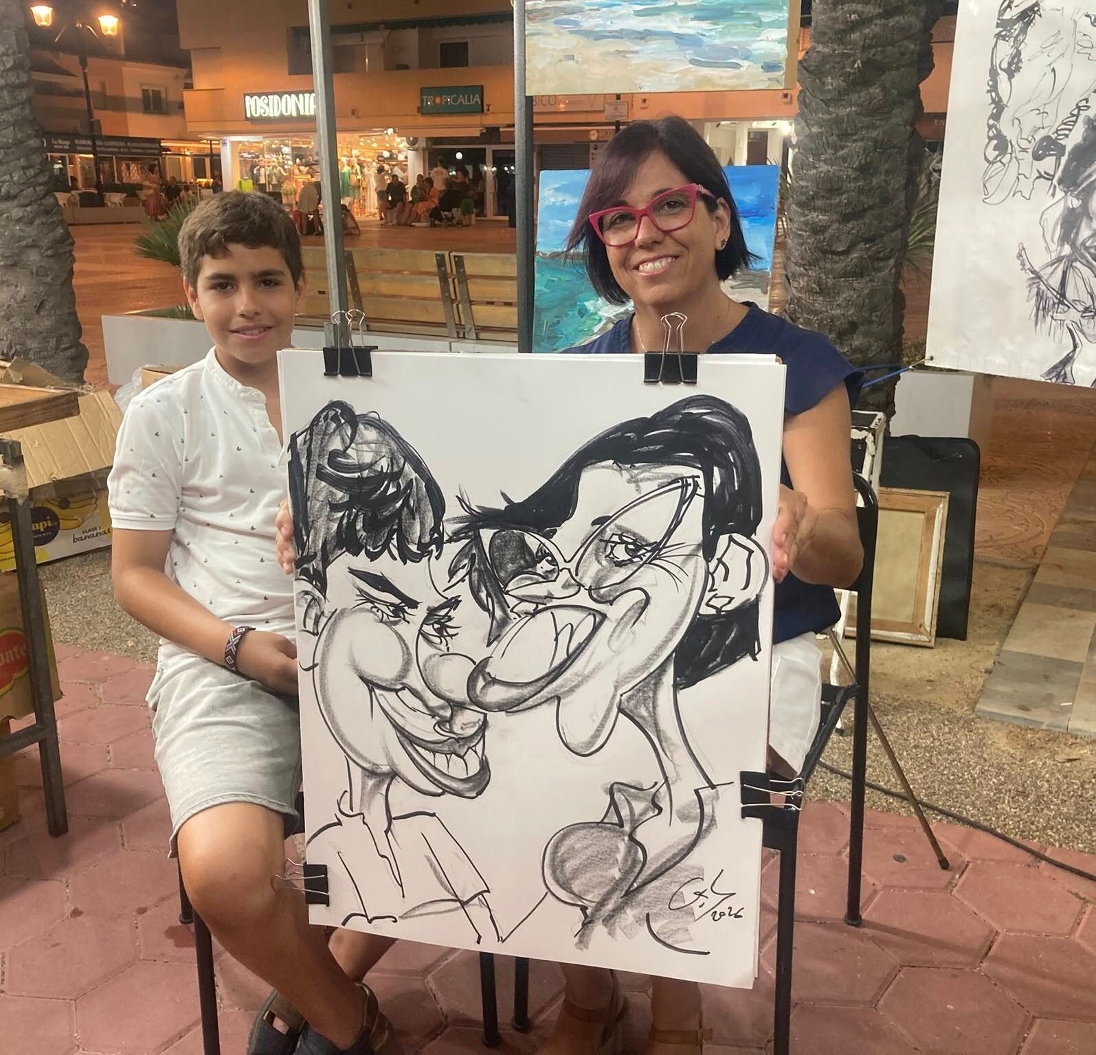 Dos personas posando con un dibujo caricaturesco en blanco y negro en una plaza al aire libre por la noche.