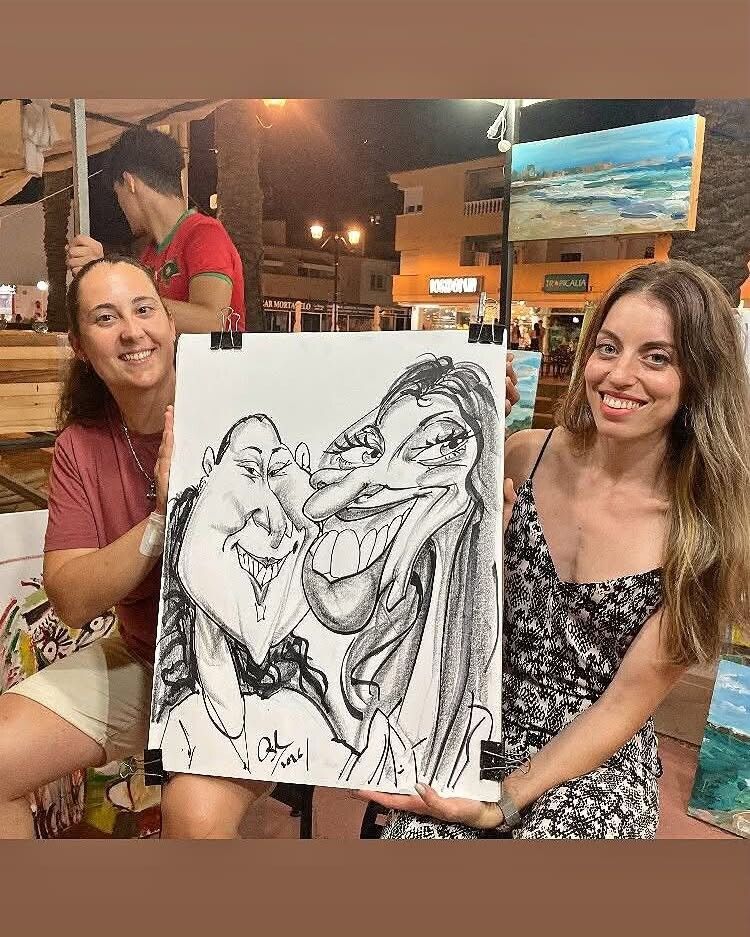 Dos mujeres sostienen un dibujo de caricatura en un local interior con iluminación cálida.