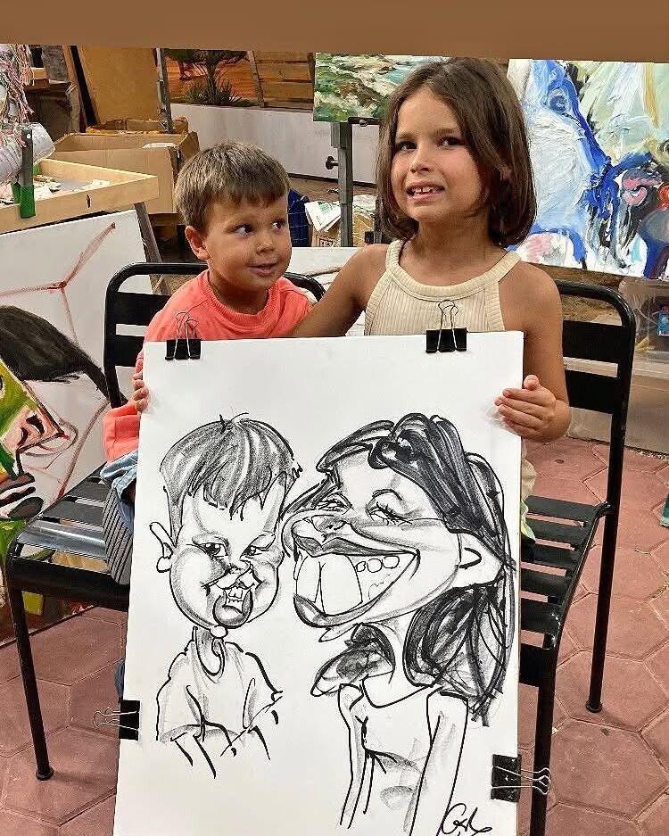 Dos niños en una feria de arte sostienen un dibujo caricaturesco de sí mismos.