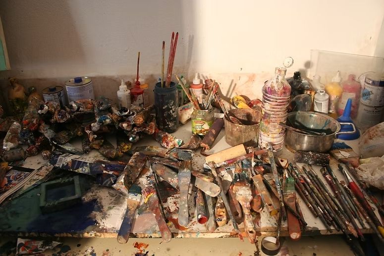 Escritorio de arte desordenado, cubierto de pinceles, tubos, frascos y coloridos materiales de pintura.