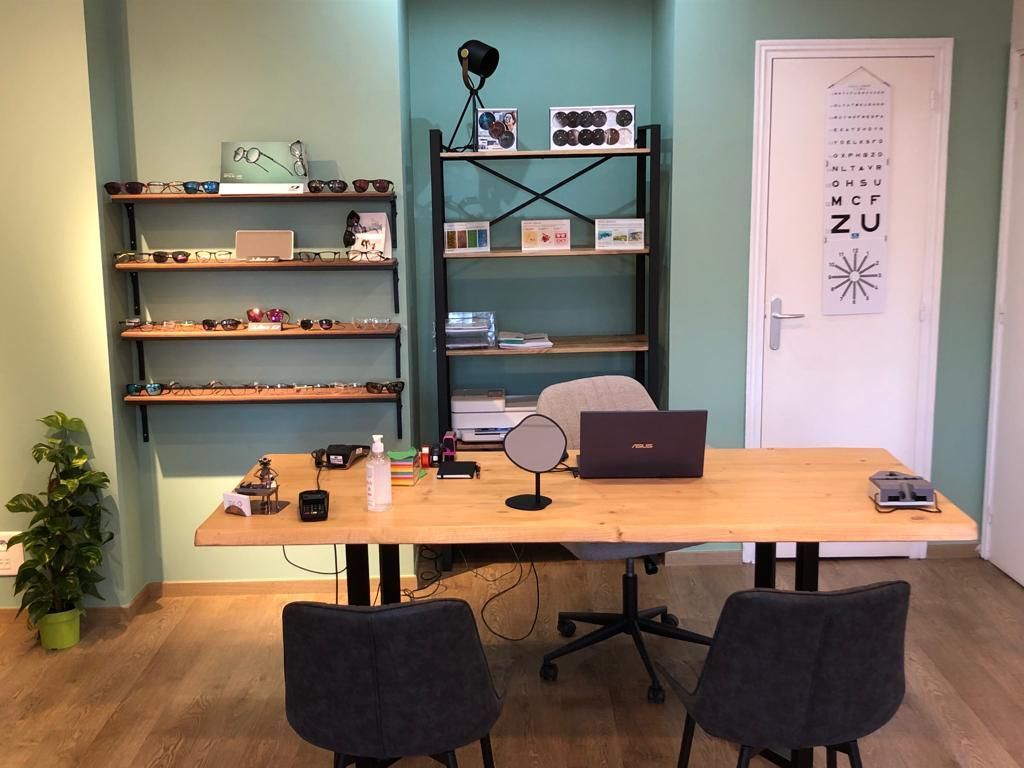 Cabinet d'optométrie : bureau en bois avec ordinateur portable, tableau d'optométrie sur la porte, étagères avec lunettes et deux chaises.