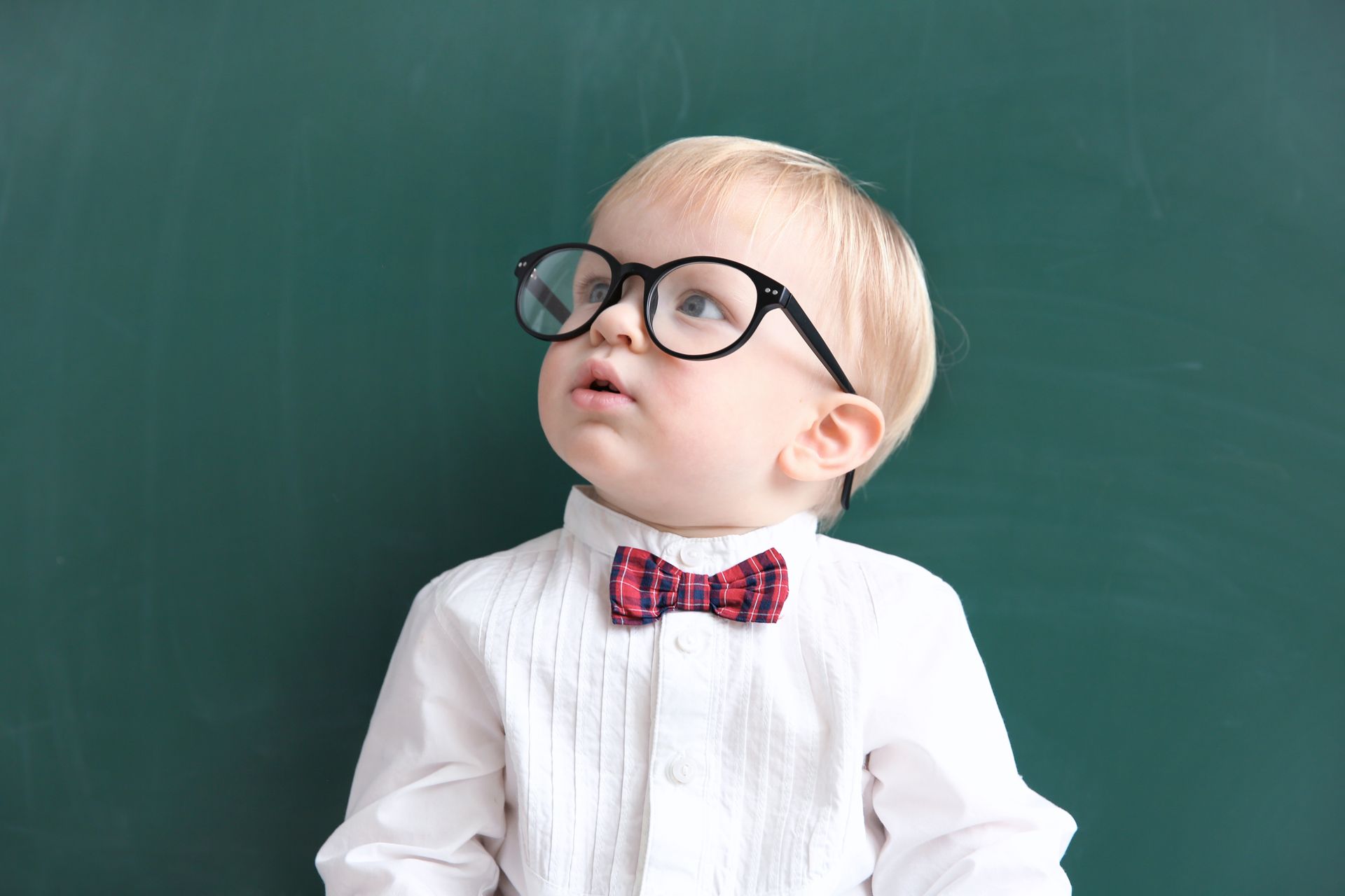 Un petit garçon blond portant des lunettes et un nœud papillon, regardant vers le haut, debout devant un tableau noir vert.