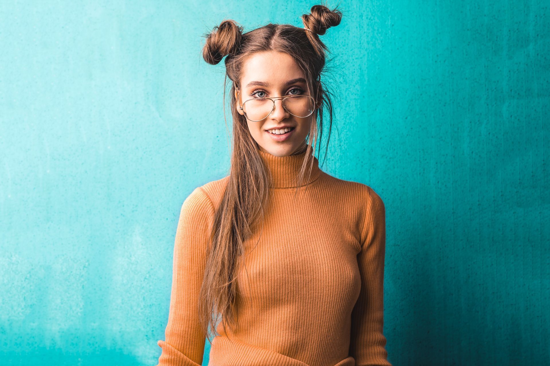 Une femme aux chignons spatiaux et portant des lunettes sourit, vêtue d'un pull à col roulé orange transparent.