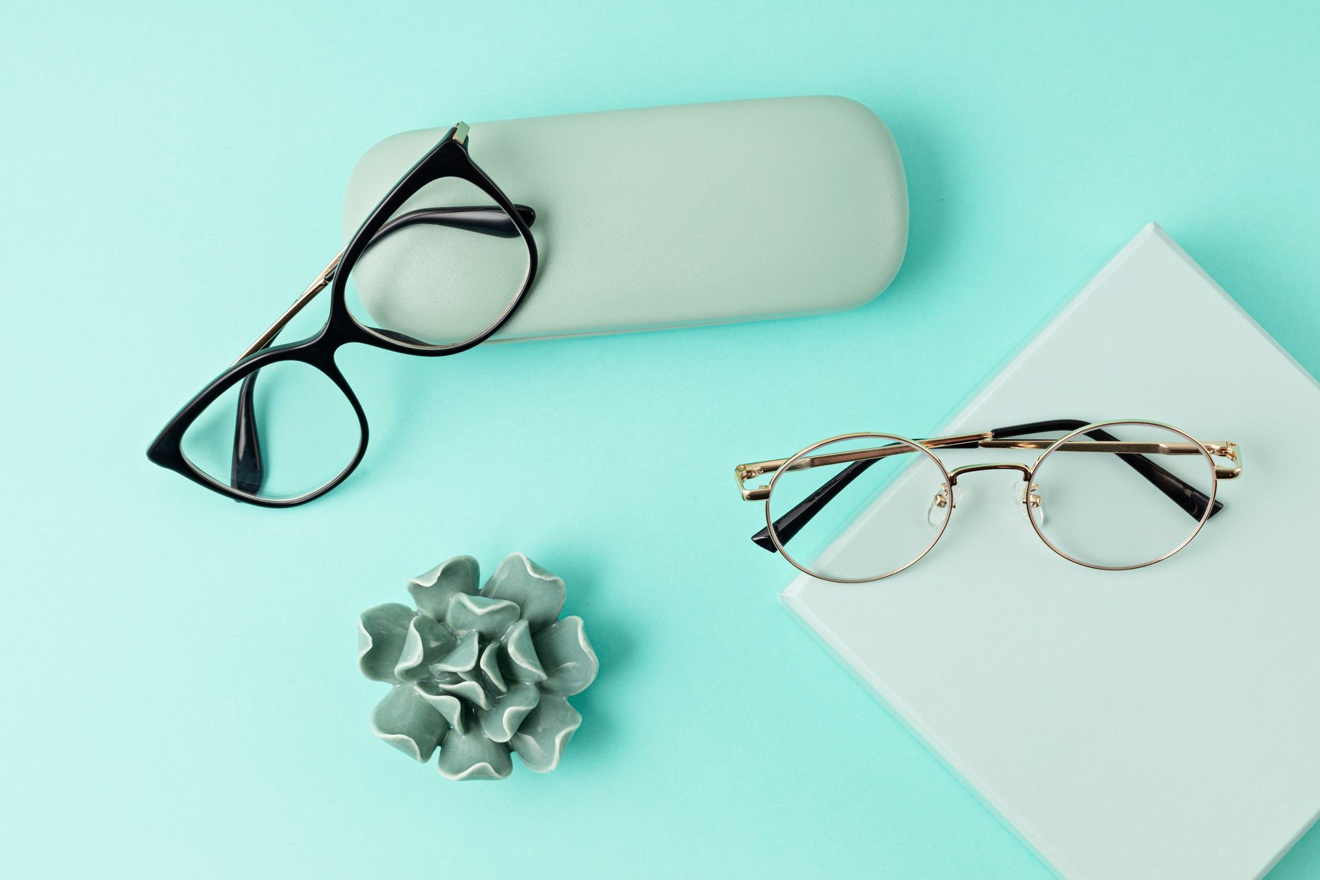 Deux paires de lunettes, un étui et une fleur décorative sur un fond turquoise.