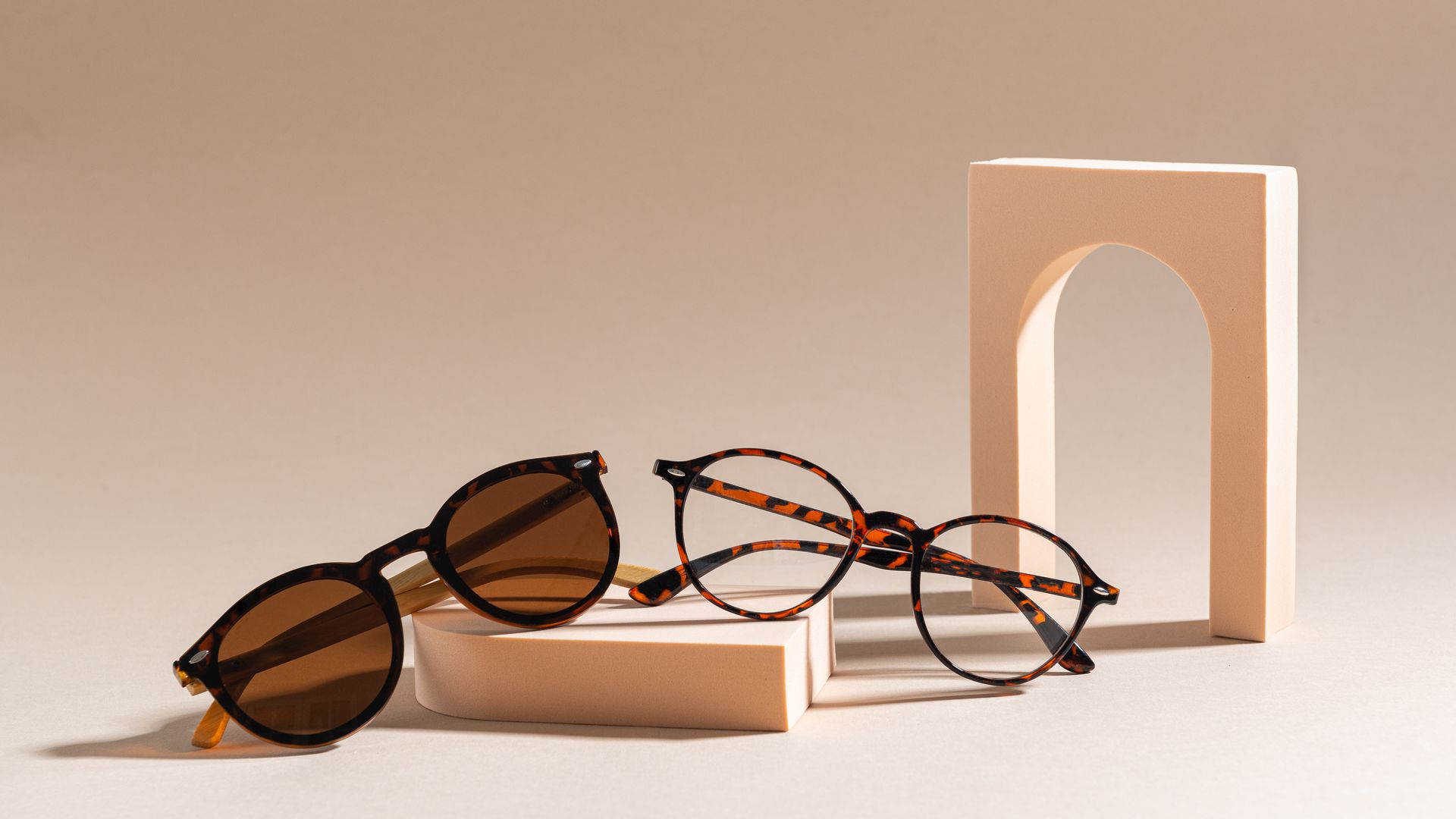 Deux paires de lunettes sur des blocs de couleur claire, avec une arche couleur pêche dans un décor aux tons neutres.