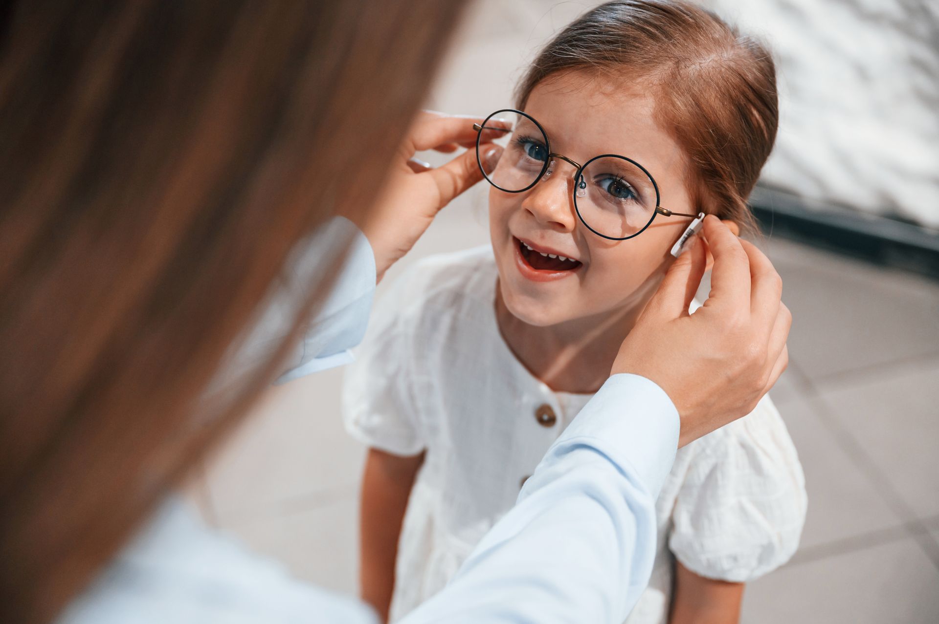 Une personne ajuste des lunettes rondes à un enfant souriant. Intérieur, robe blanche, peau claire.