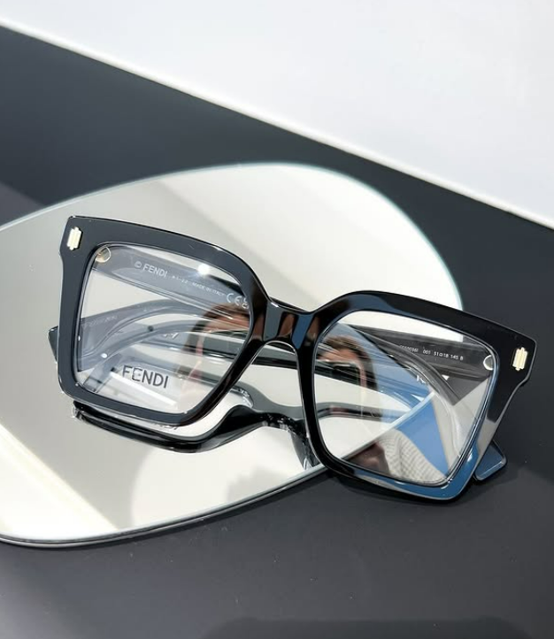 Lunettes Fendi noires sur fond réfléchissant. La monture est épaisse, carrée et brillante.