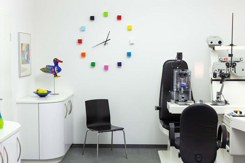 Untersuchungszimmer eines Augenarztes: weiße Wände, bunte Uhr, Stuhl, Untersuchungsgeräte für die Augenuntersuchung und eine Statue auf dem Schrank.