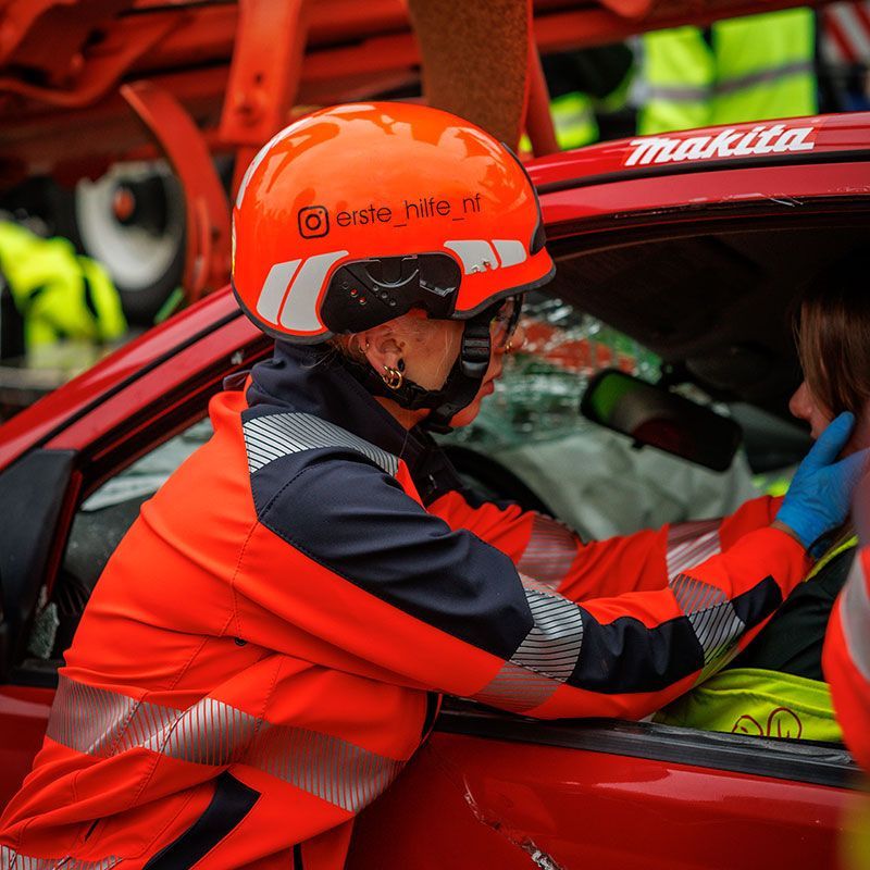 Ein Rettungshelfer in orangefarbener Schutzkleidung leistet einem Fahrer in einem roten Auto nach einem Unfall Hilfe.
