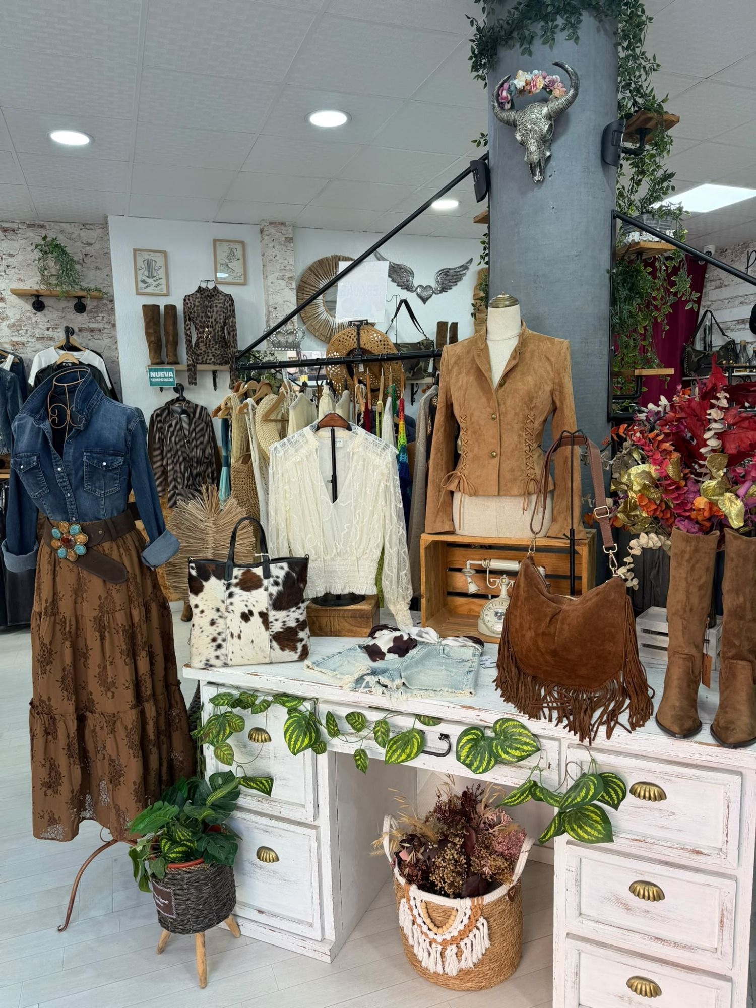 Exhibidor de una boutique de ropa con vestidos vintage, abrigos, botas y decoración floral alrededor de una cómoda blanca
