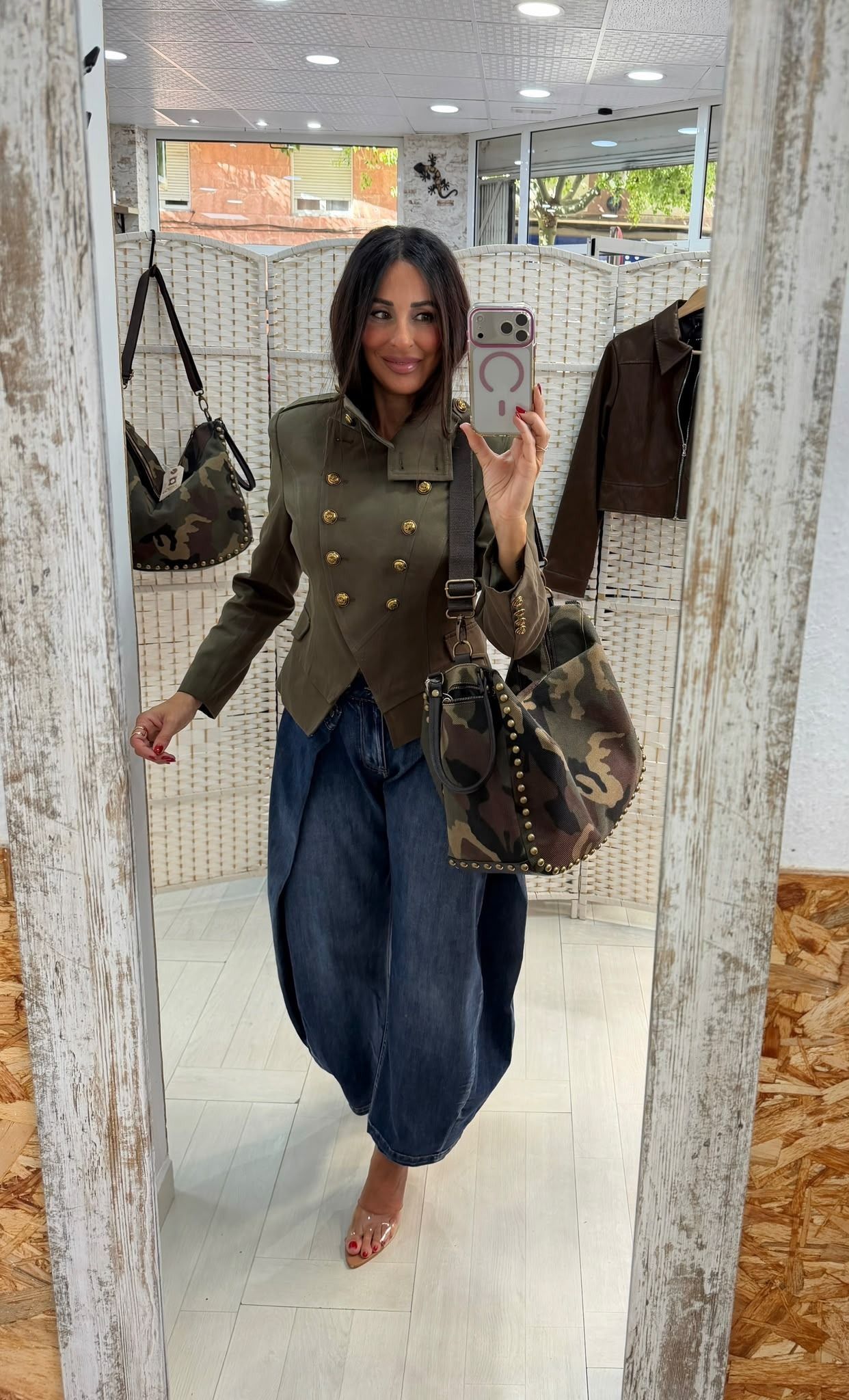 Mujer haciéndose un selfi frente al espejo en una tienda de ropa, con un top verde oliva y unos vaqueros azules, y llevando un bolso marrón.