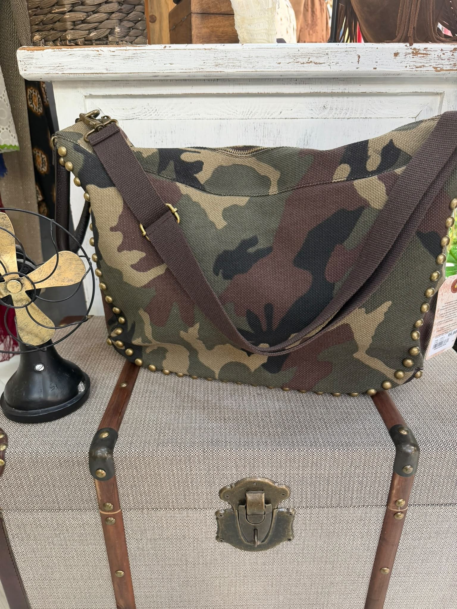 Bolso de camuflaje sobre una silla de mimbre frente a una repisa blanca, con un farol en el suelo cerca