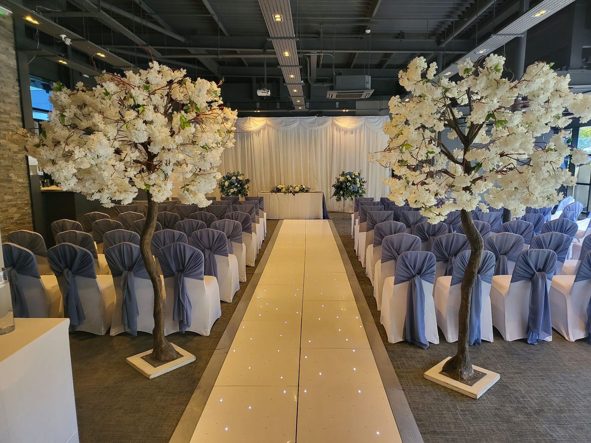 Wedding ceremony setup with rows of blue-sashed chairs, white aisle, and cherry blossom trees.