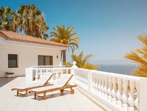 Dos sillas de salón en una terraza blanca con vista al mar, palmeras al fondo, día soleado.