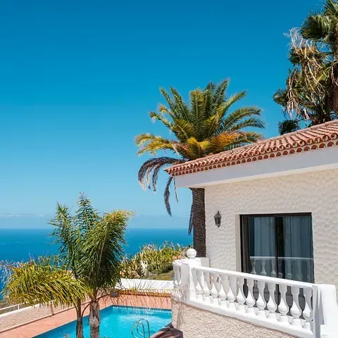 Una villa blanca con piscina, con vistas a un océano azul bajo un cielo azul brillante, con palmeras.