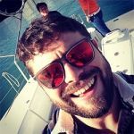 Hombre con barba sonriendo, con gafas de sol con lentes rojas, en un barco con otra persona al fondo.