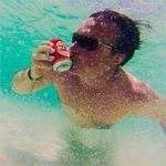 Hombre con gafas de sol bebiendo de una lata bajo el agua en aguas turquesas claras.