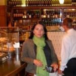 Una mujer con una bufanda verde se encuentra junto al mostrador de un café con botellas de vino en el fondo.