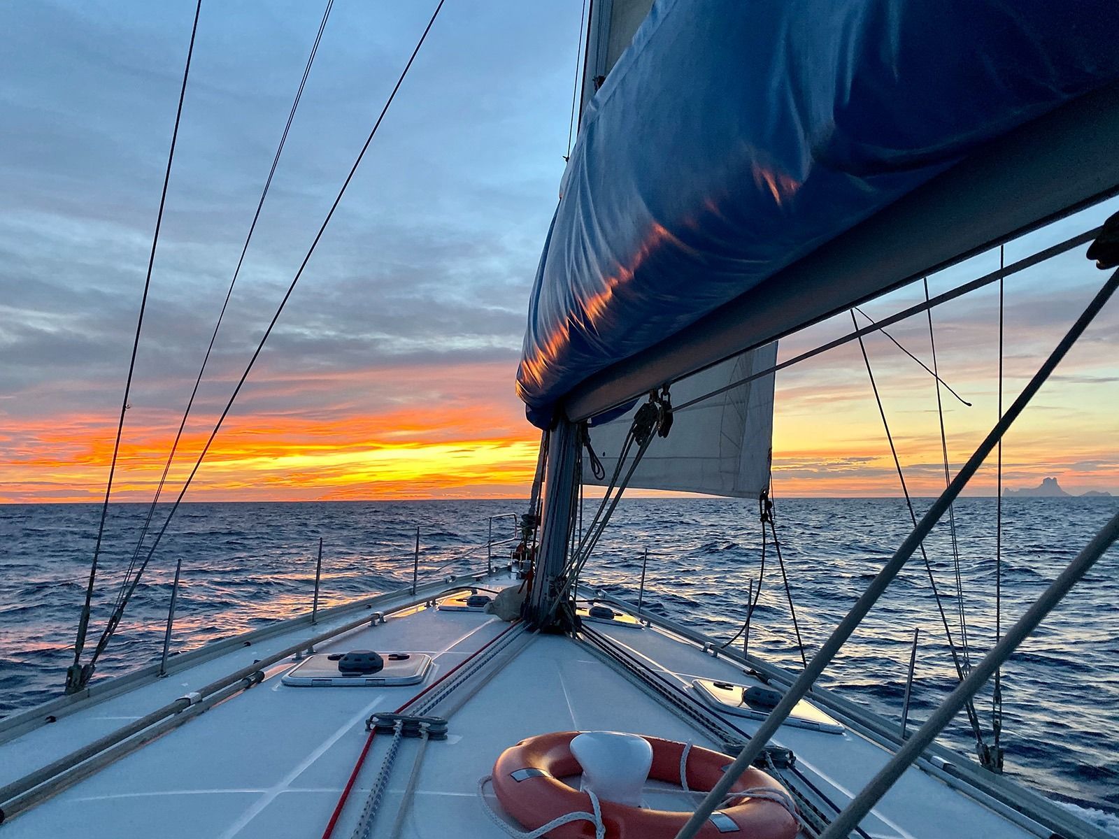 Velero navegando en el océano durante la puesta de sol, cielo naranja y azul, salvavidas visible en la cubierta.