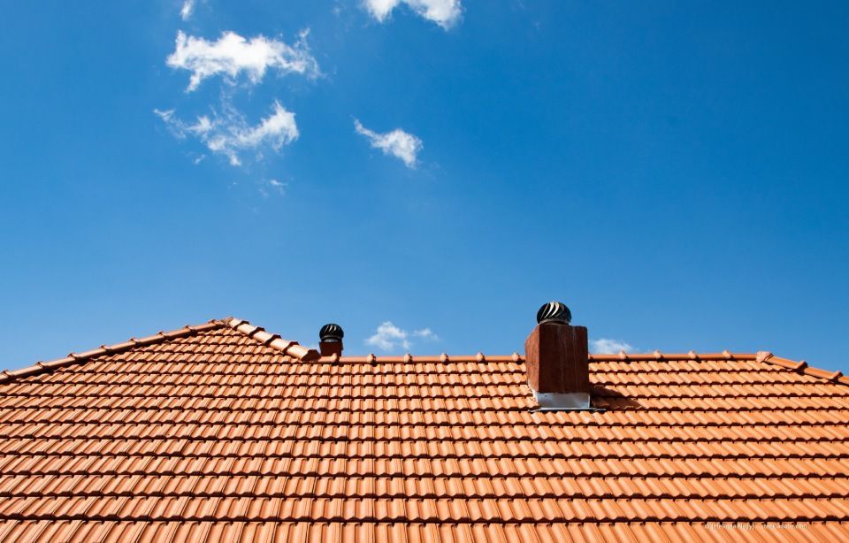Rotes Ziegeldach mit zwei Lüftungsöffnungen und einem gemauerten Schornstein vor einem strahlend blauen Himmel mit vereinzelten Wolken.