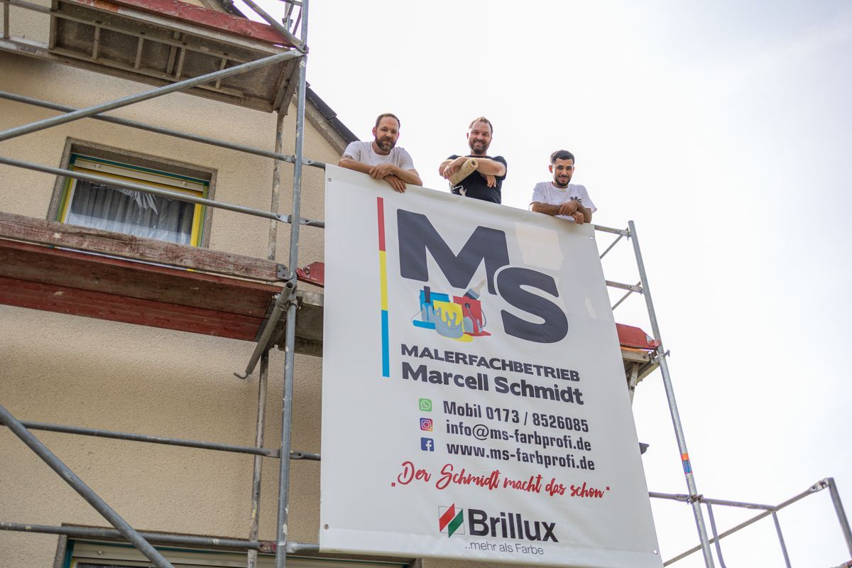 Drei Männer stehen auf einem Gerüst und halten ein Schild mit der Aufschrift „Ms.“