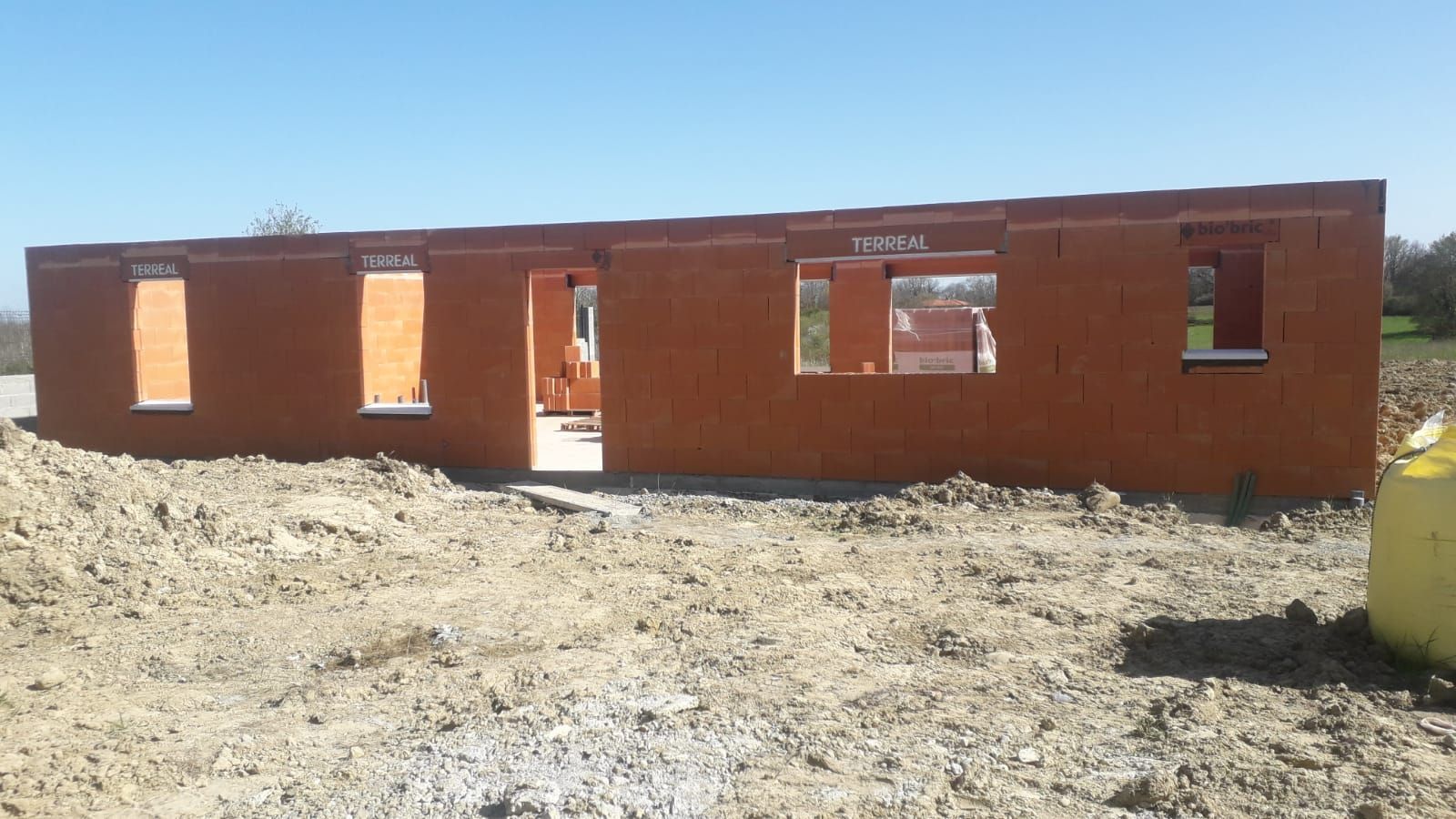 Maison à ossature bois en construction sur un terrain sec, avec des conteneurs à déchets verts et un ciel bleu dégagé.