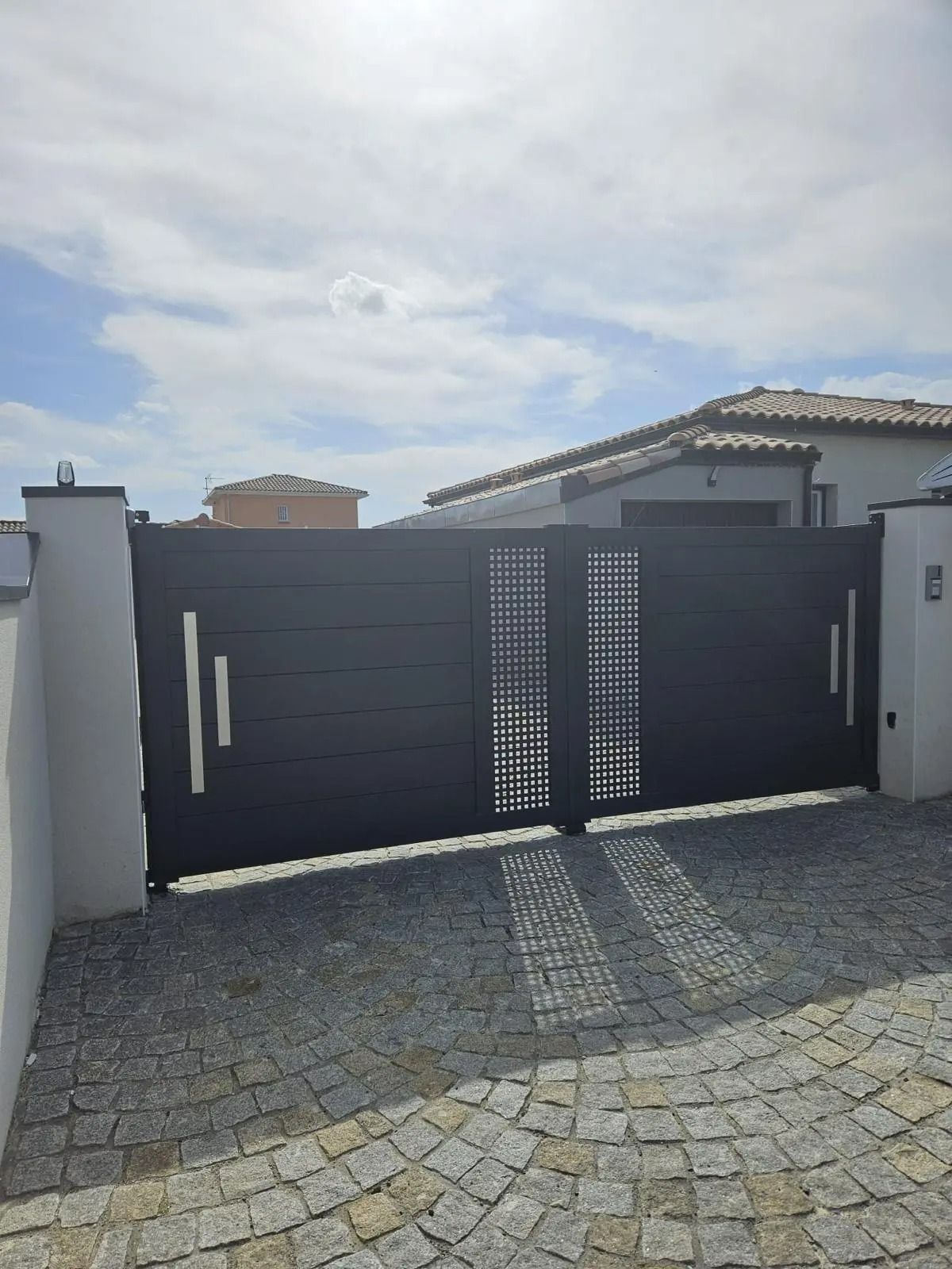 Portail d'allée en métal gris foncé avec poteaux blancs et pavage en pavés devant une maison
