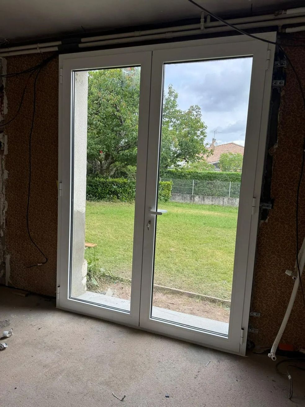 Des portes-fenêtres blanches dans une pièce inachevée, donnant sur une cour verdoyante et une haie à l'extérieur.
