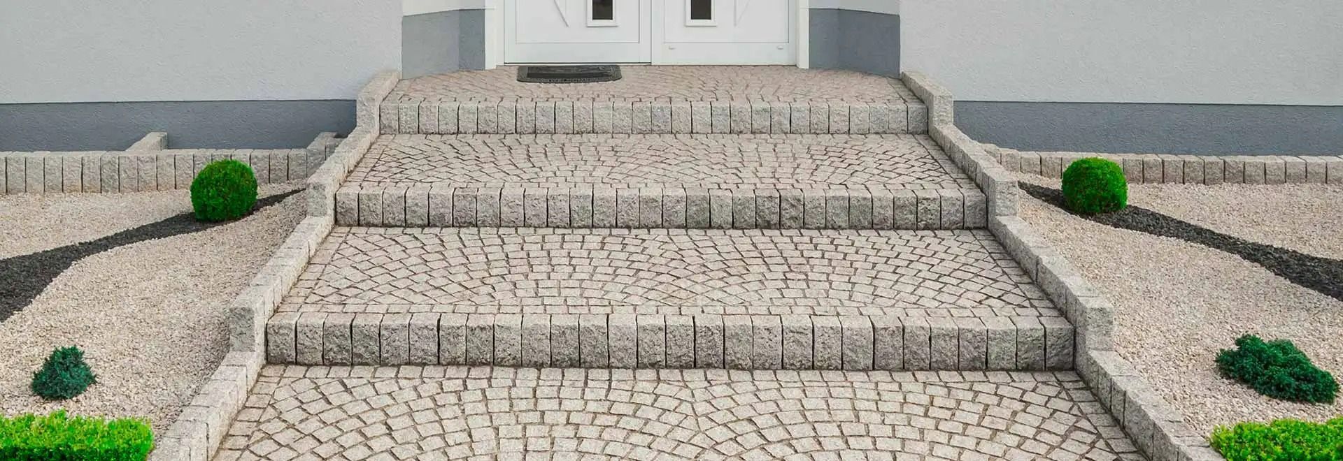 Des marches en pierre mènent à un bâtiment blanc, bordé de petits arbustes verts de part et d'autre.