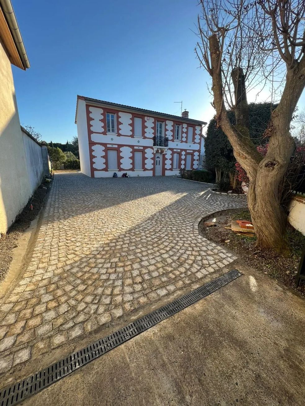 Cour pavée avec un bâtiment rouge et blanc sous un ciel bleu clair, bordée d'arbres et d'un mur jaune
