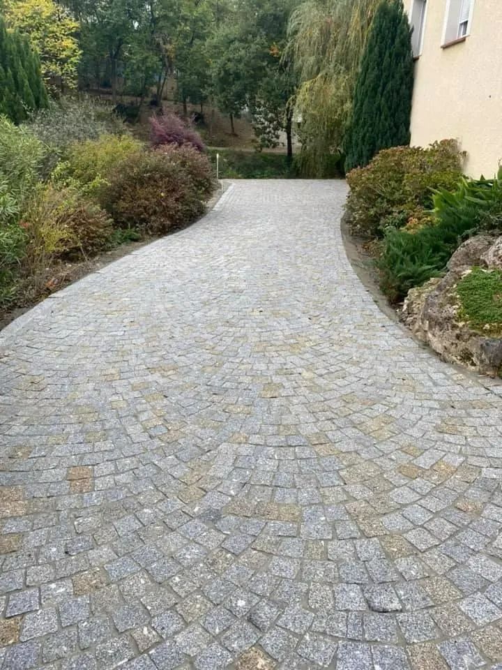 Allée de jardin pavée et sinueuse longeant une maison beige, bordée d'arbustes et d'arbres