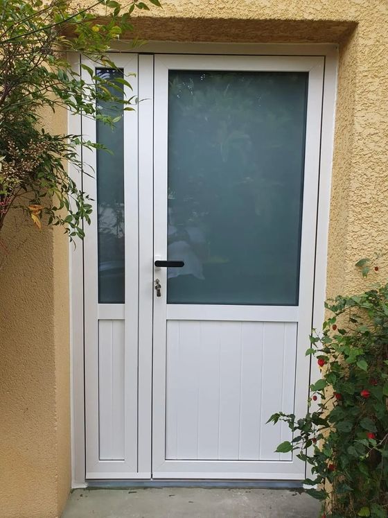 Porte d'entrée blanche avec panneau en verre dépoli, encadrée de stuc beige et de verdure.