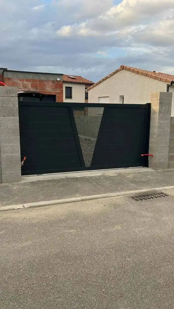 Portail en métal noir devant une maison et un muret dans une rue résidentielle sous un ciel nuageux