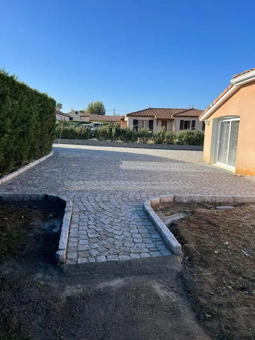 Cour pavée jouxtant une maison beige et des haies, sous un ciel bleu azur.