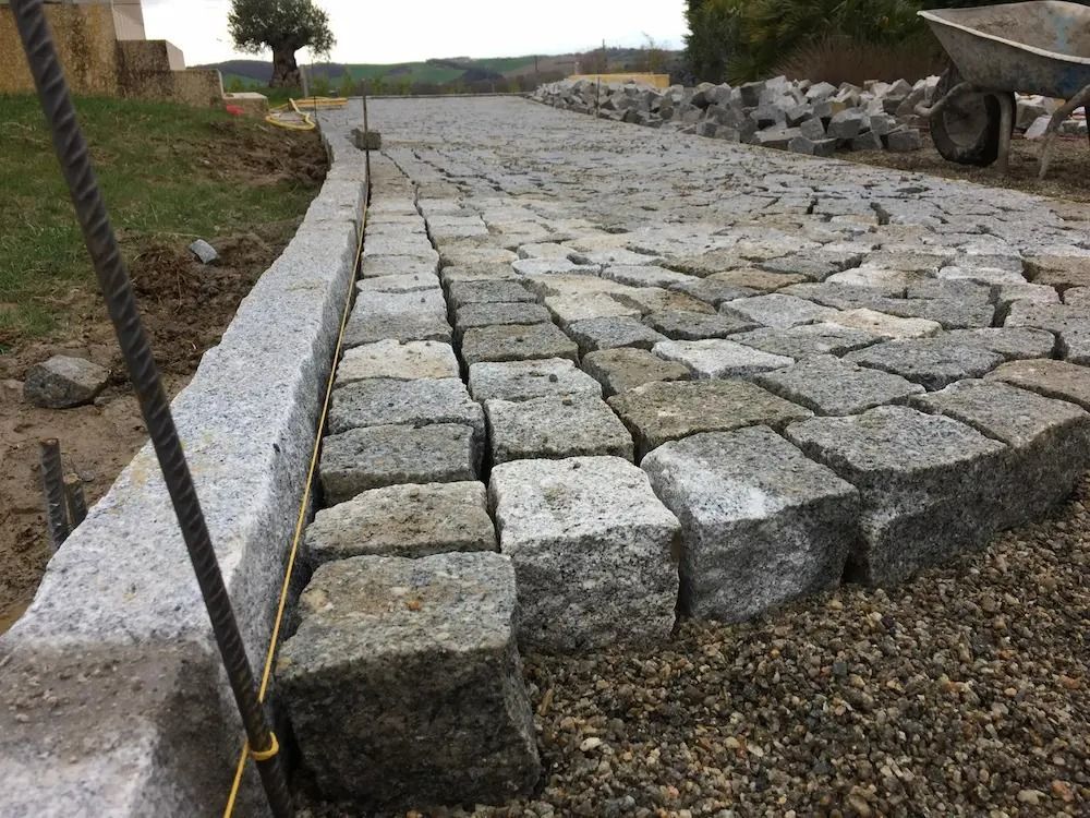 Allée en pierre en construction, avec de grandes dalles grises, bordée d'un chemin de gravier et d'une bordure rocheuse.