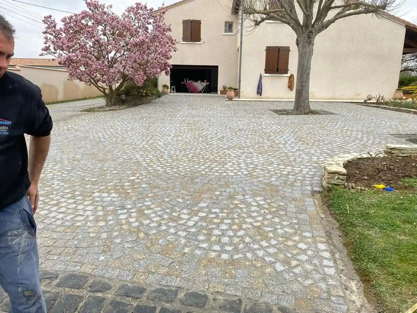 Allée de gravier menant à une maison blanche, avec un arbre en fleurs roses et une personne partiellement visible sur la gauche.