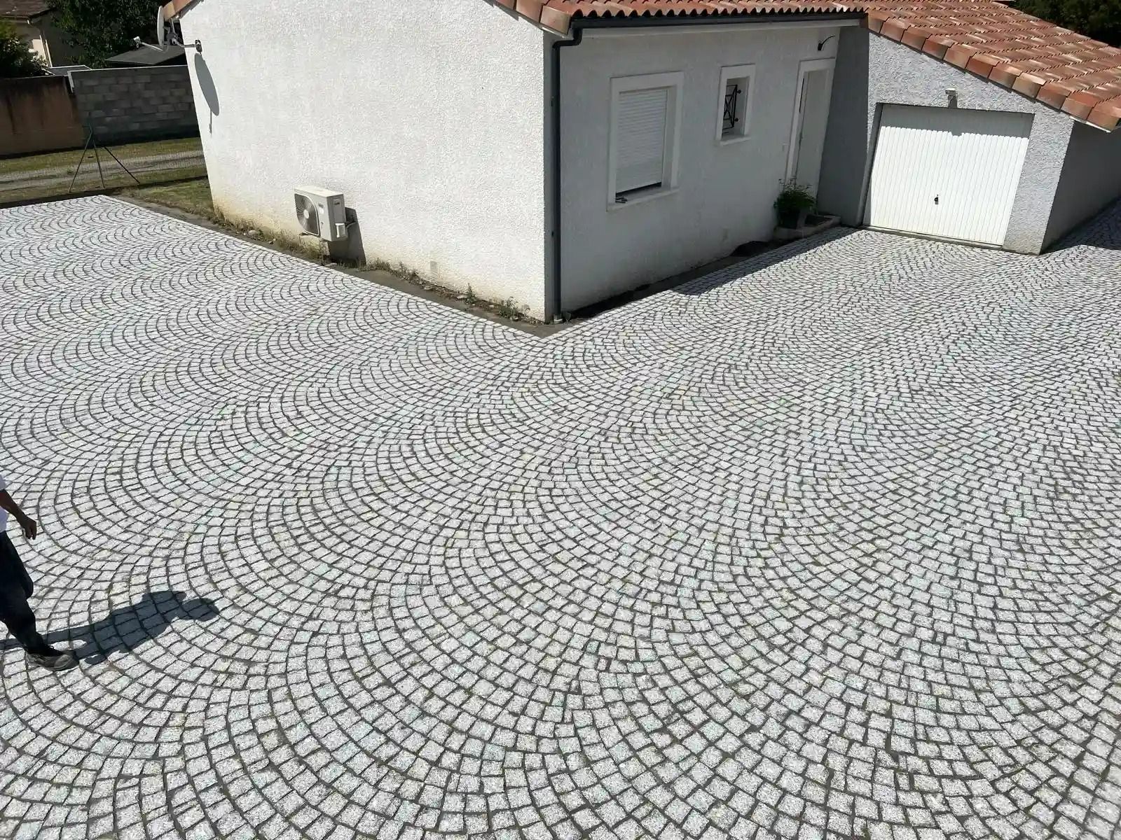 Cour pavée bordée de bâtiments blancs aux toits de tuiles rouges, avec l'ombre d'une personne sur le bord gauche.