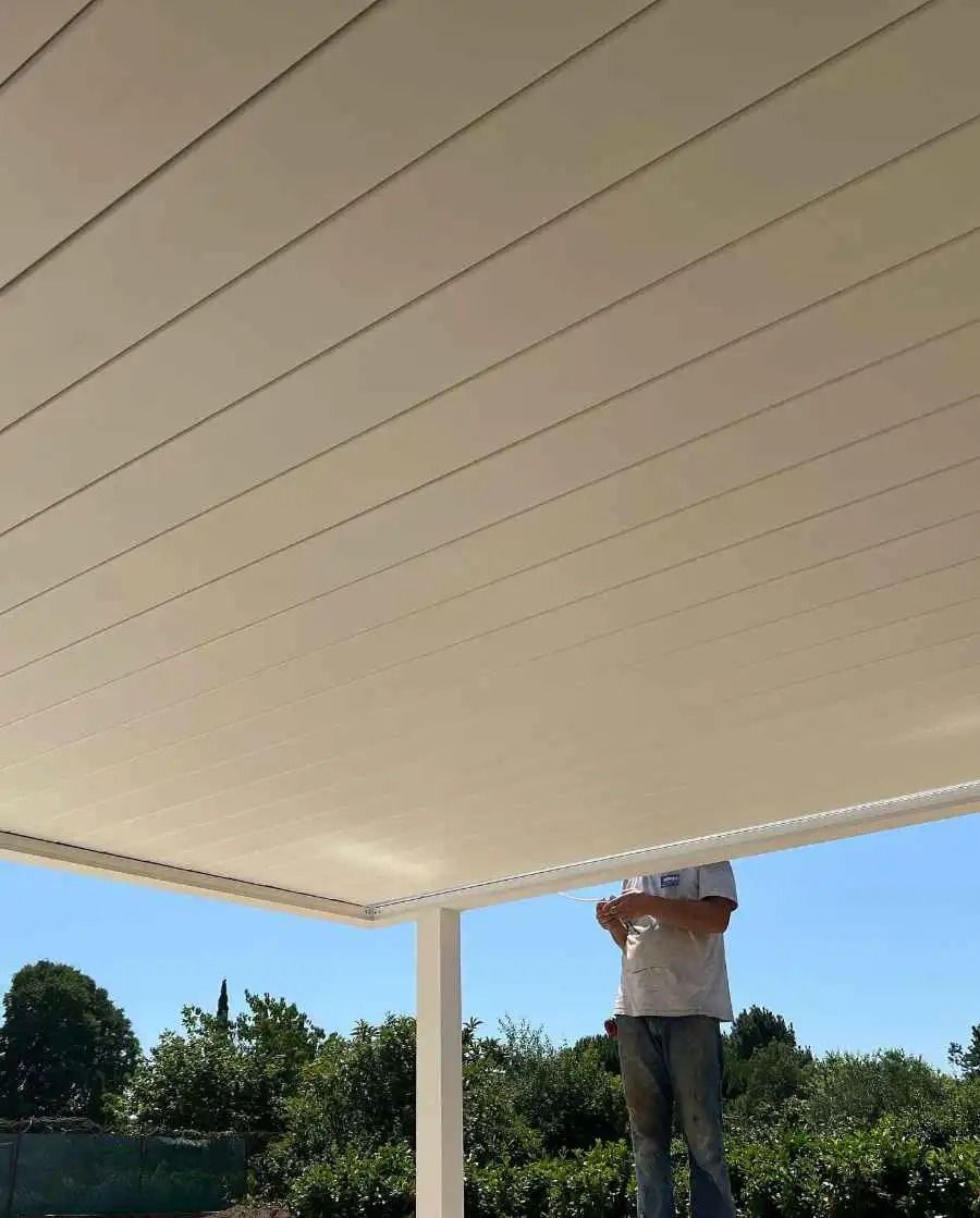 Une personne debout sur une terrasse sous un toit blanc, tendant le bras pour ajuster quelque chose près du bord.