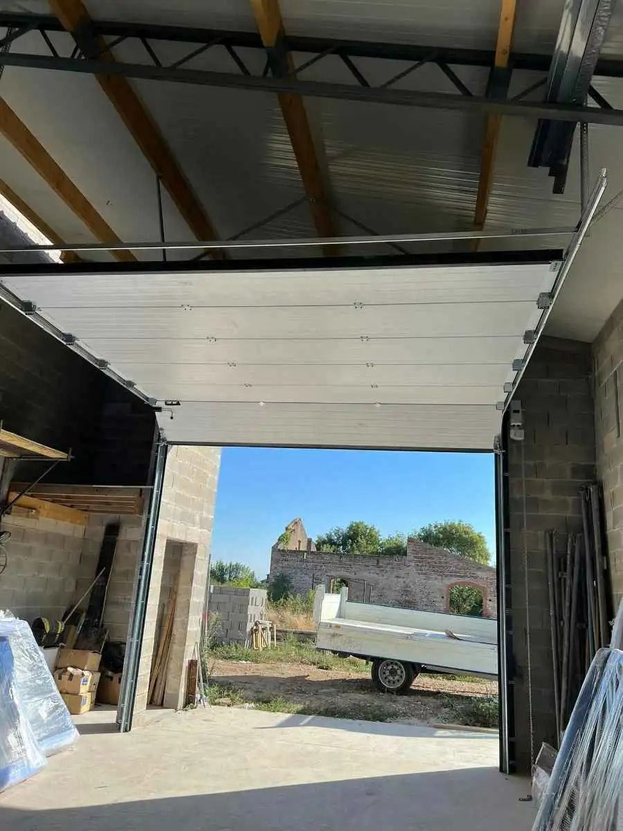 À l'intérieur d'un garage, une porte sectionnelle ouverte encadre une petite remorque garée à l'extérieur par une journée ensoleillée.