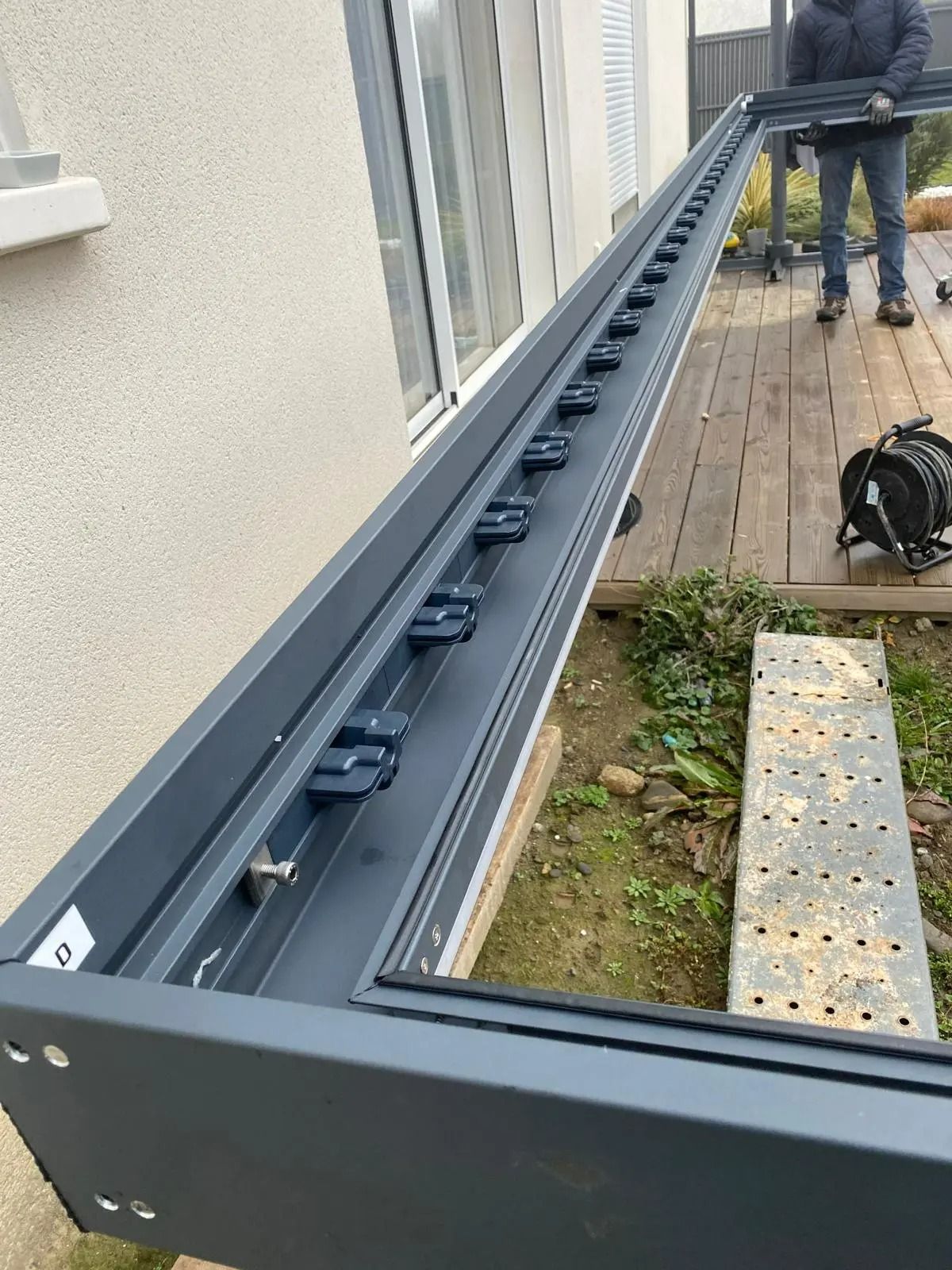 Jardinière en métal gris le long d'un mur d'immeuble, avec des matériaux de construction et un ouvrier en arrière-plan.