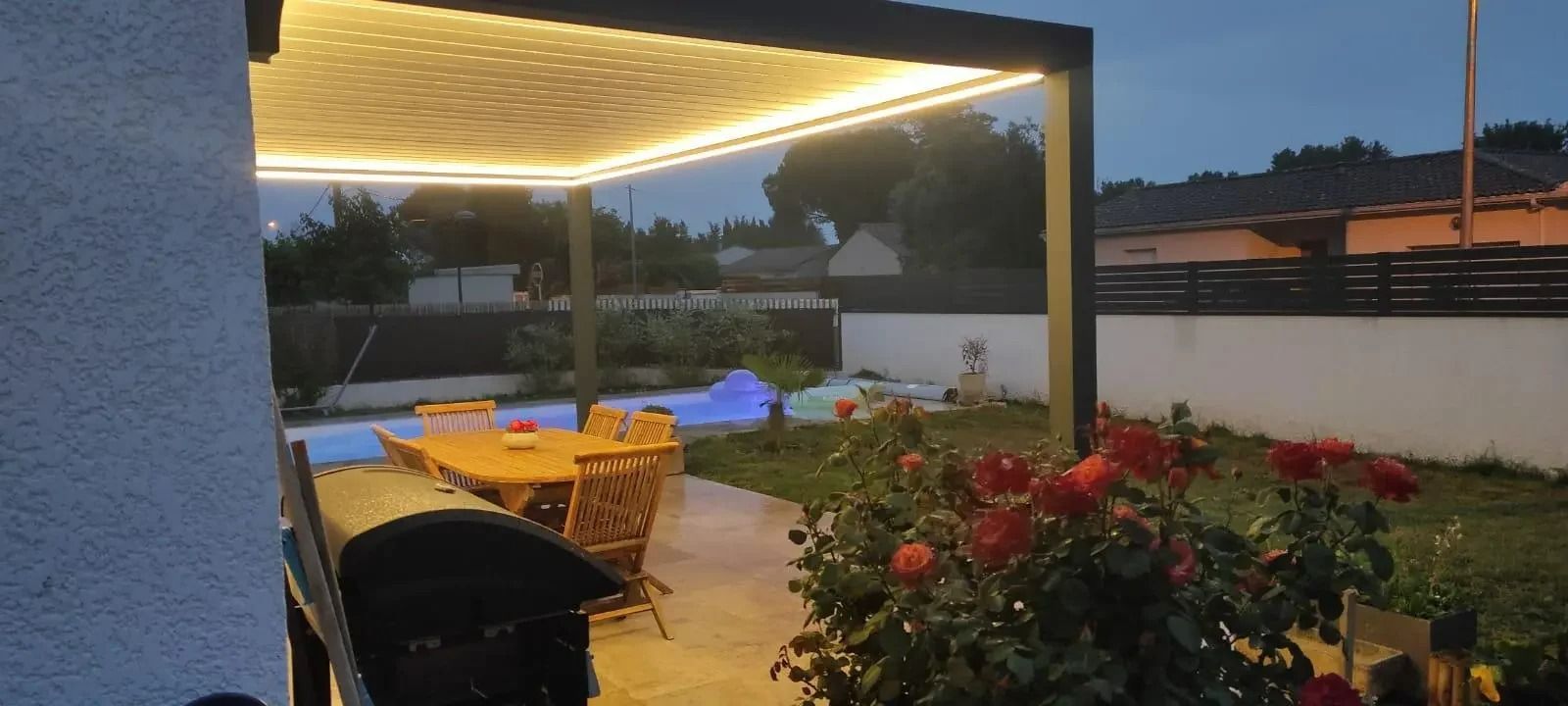 Terrasse au crépuscule, éclairée par des lumières chaudes au plafond, table à manger, chaises et fleurs rouges près d'un muret.