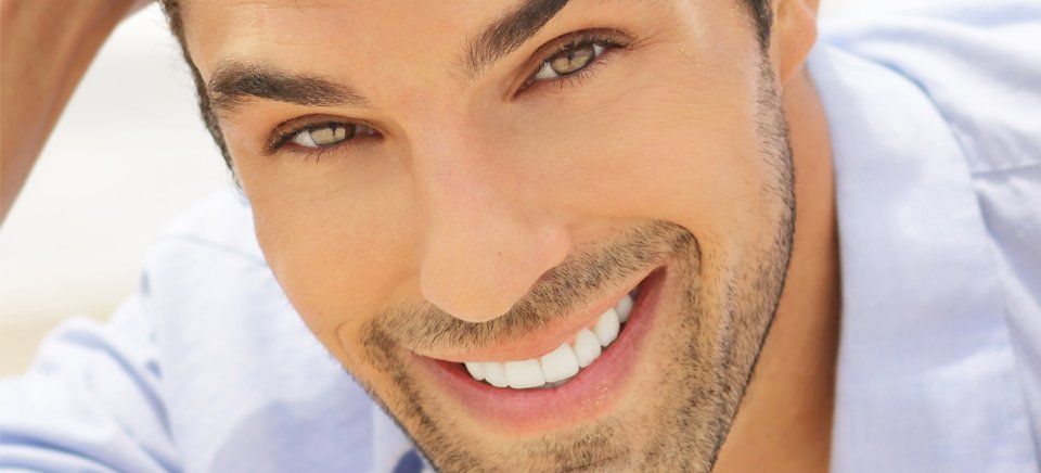 Un hombre de ojos marrones sonríe, viste una camisa azul claro.