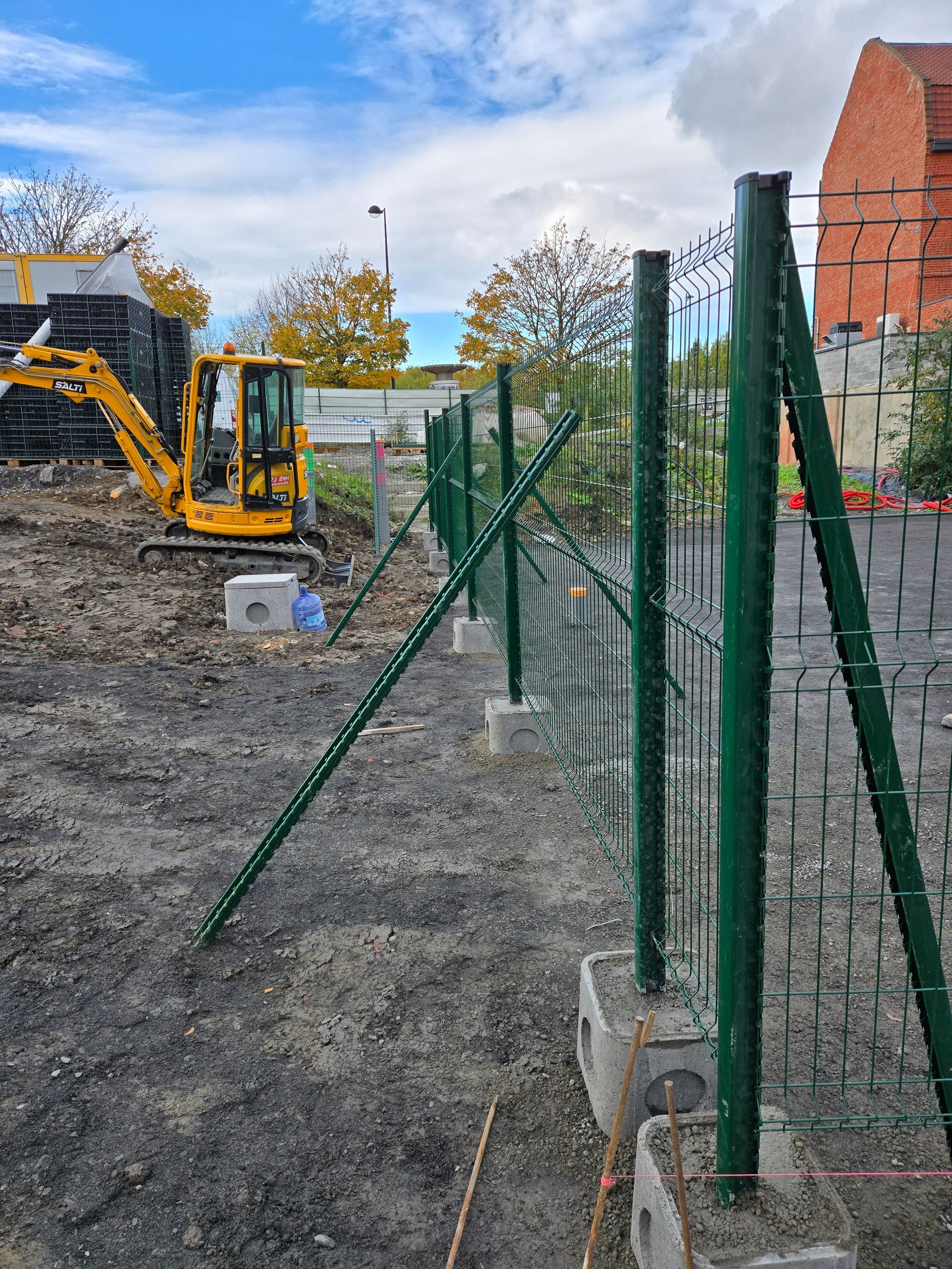 Clôture de chantier en métal vert. Une pelleteuse jaune est visible en arrière-plan.