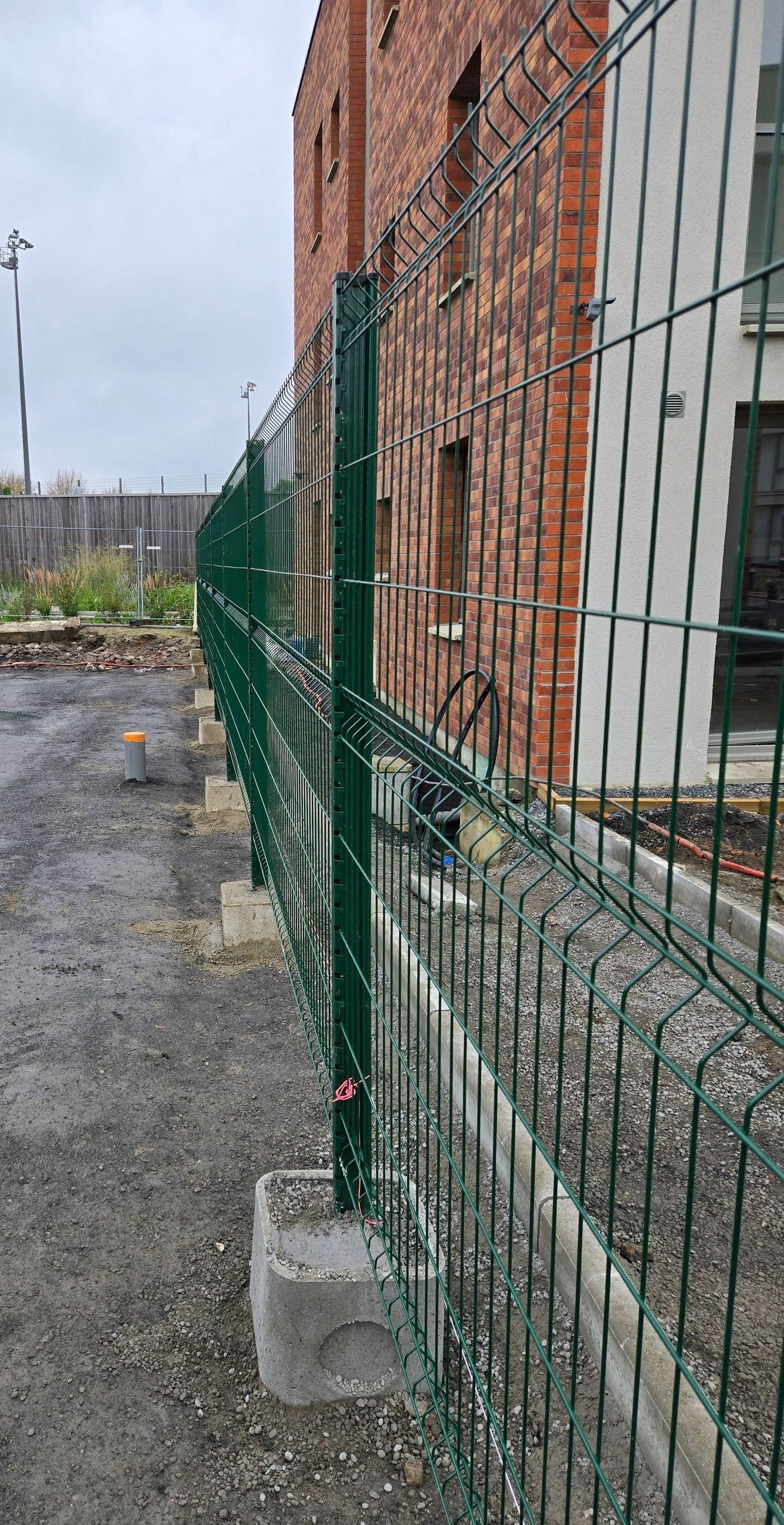 Une clôture en fil de fer vert surmontée de barbelés longe un bâtiment en briques et un chemin de gravier.