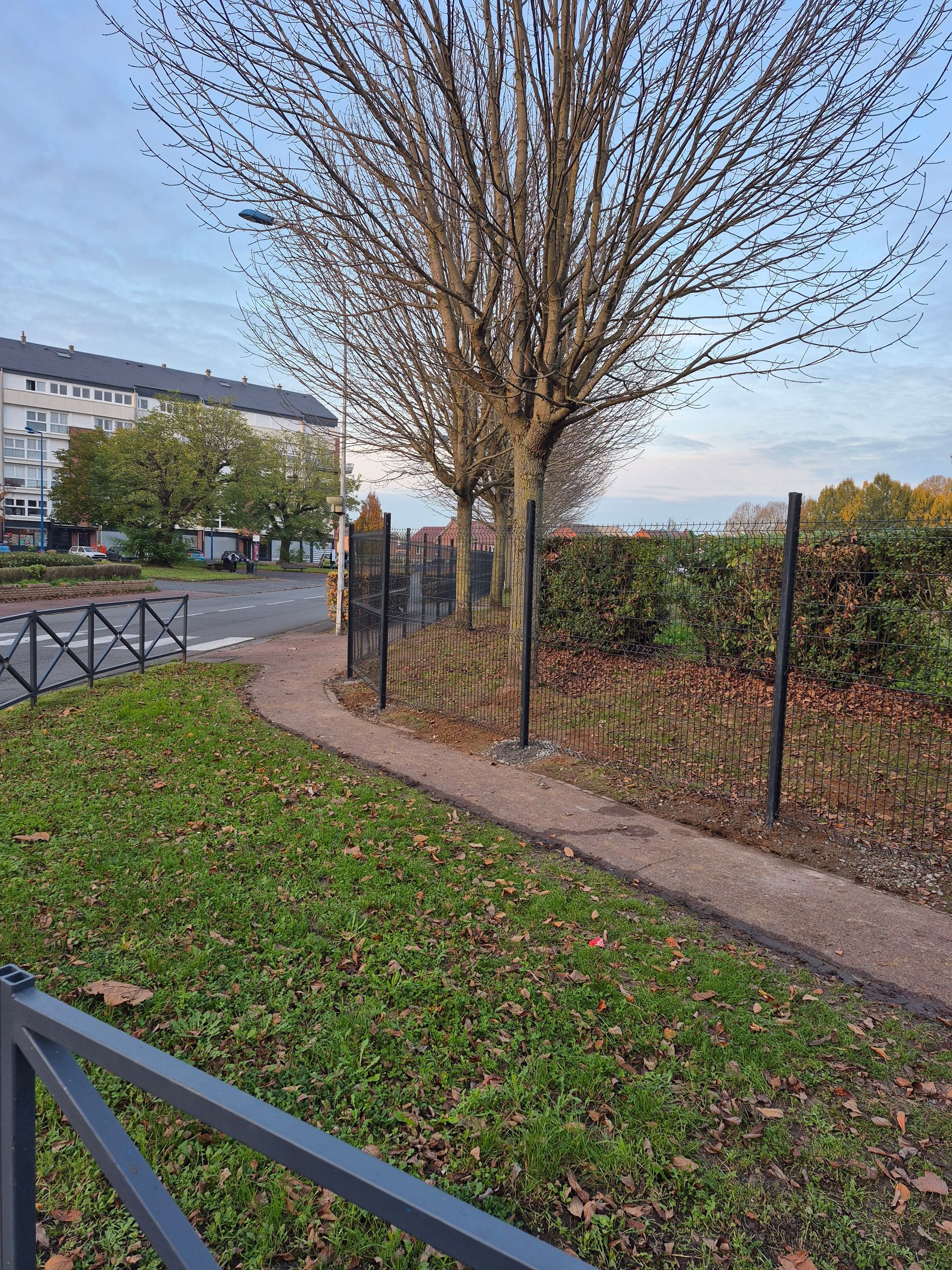 Un chemin de gravier mène à un portail offrant une vue sur une rue et des bâtiments ; une zone herbeuse et des arbres bordent le chemin de chaque côté.
