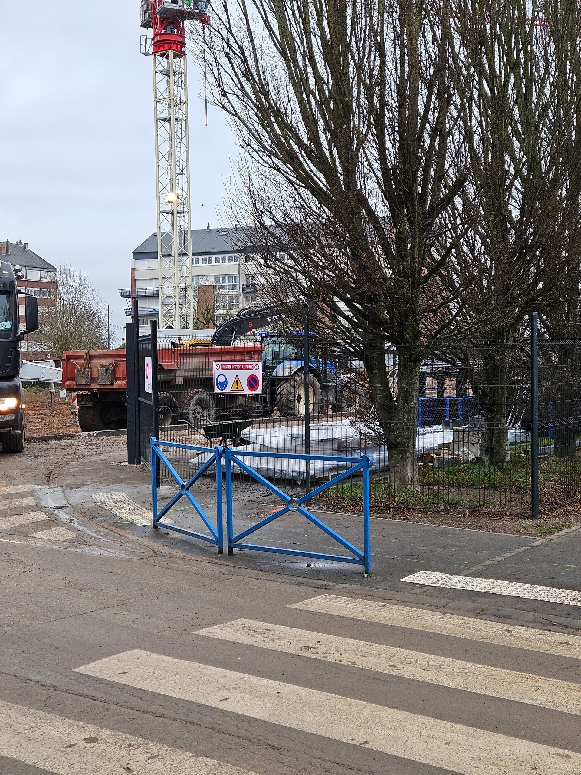 Chantier de construction avec grue, camion et portail bleu sur un passage piéton.