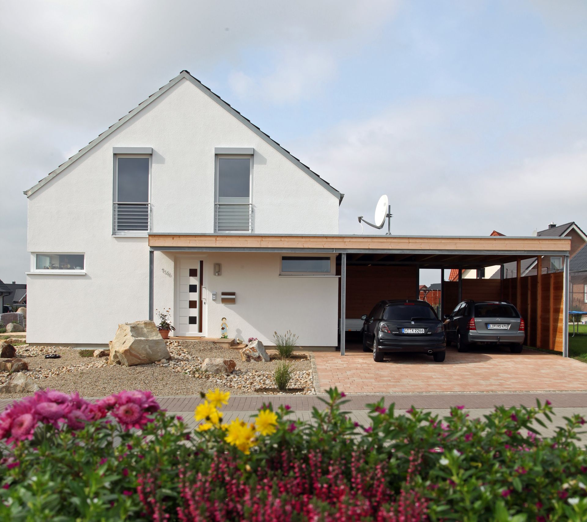 Maison blanche avec abri voiture, deux voitures garées, jardin fleuri, ciel bleu.