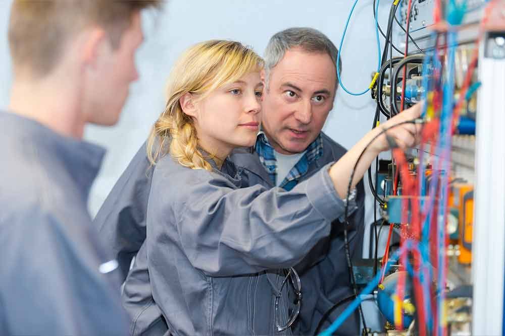 Studenten und Dozent untersuchen elektrische Leitungen in einem Labor und zeigen auf Bauteile.