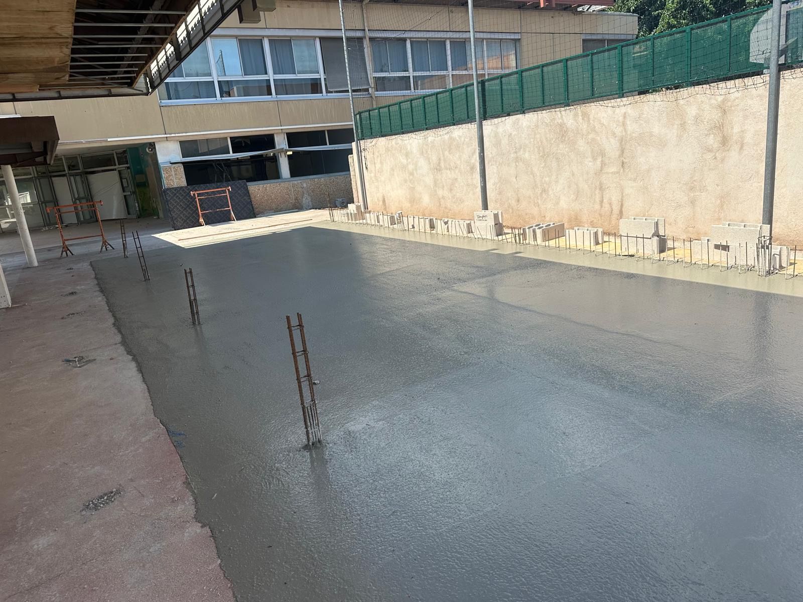 Route en béton fraîchement coulé avec des poteaux métalliques, un mur et un bâtiment en arrière-plan.