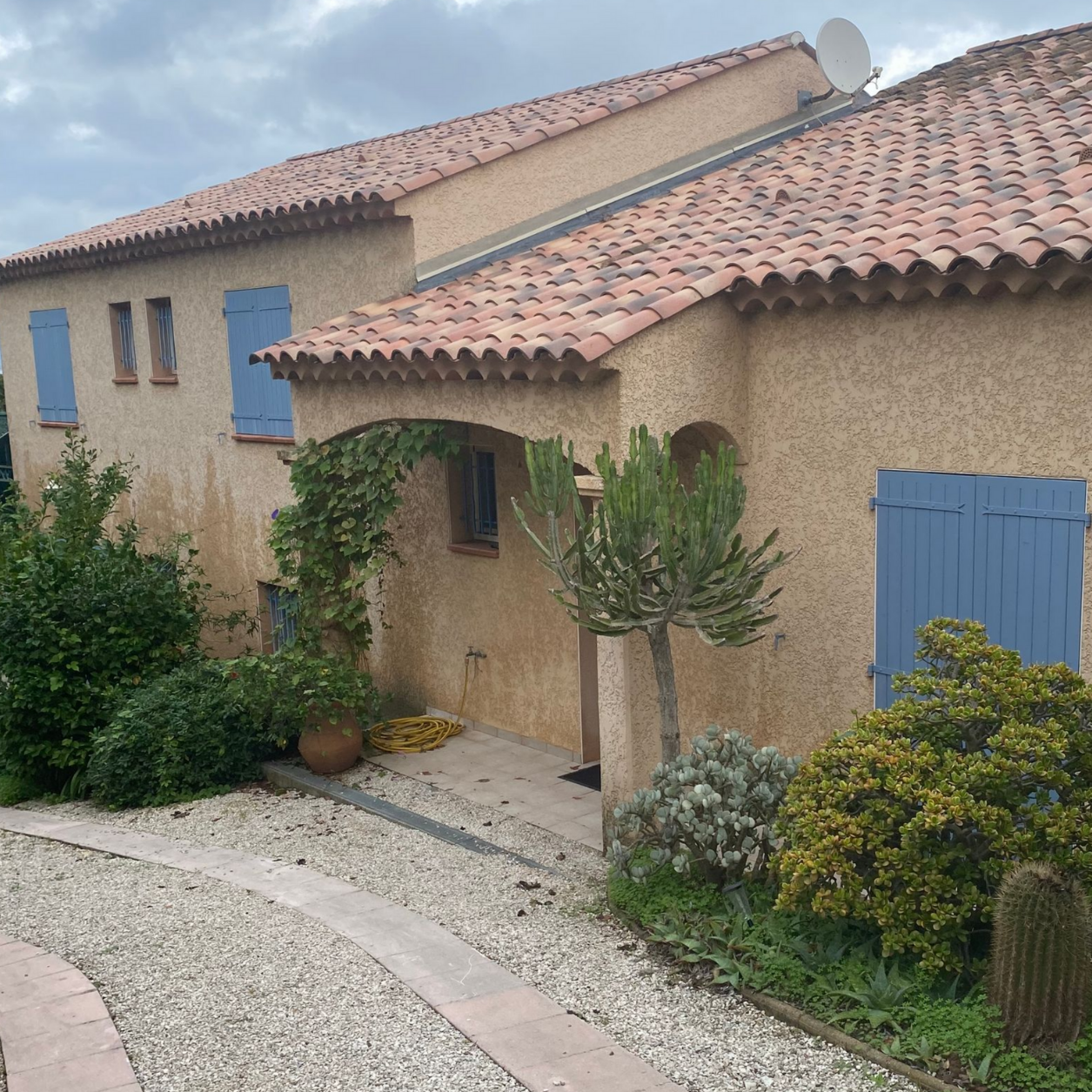Maison en stuc beige avec toit de tuiles rouges, volets bleus et allée de gravier.